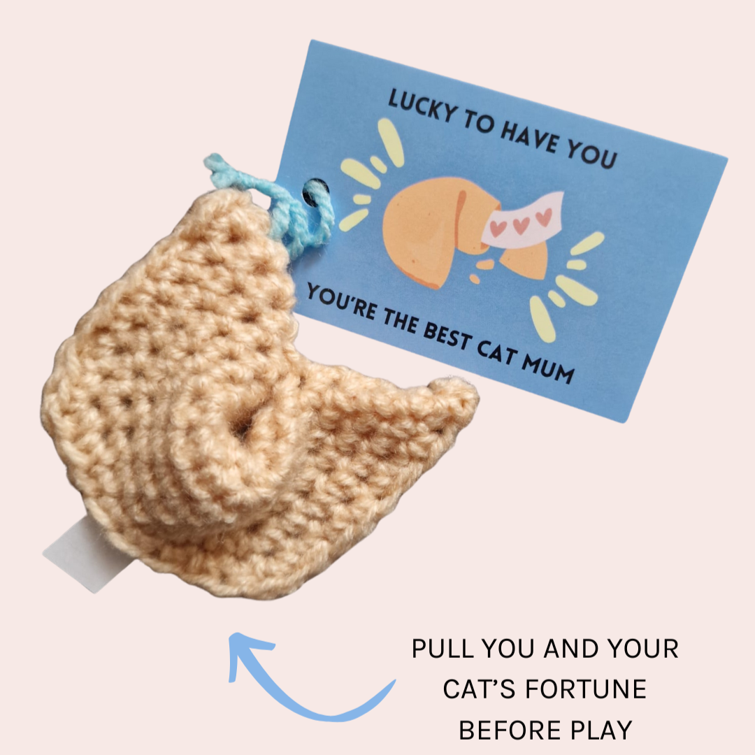 Crocheted cat toy shaped like a fortune cookie with a card and text on a light pink background