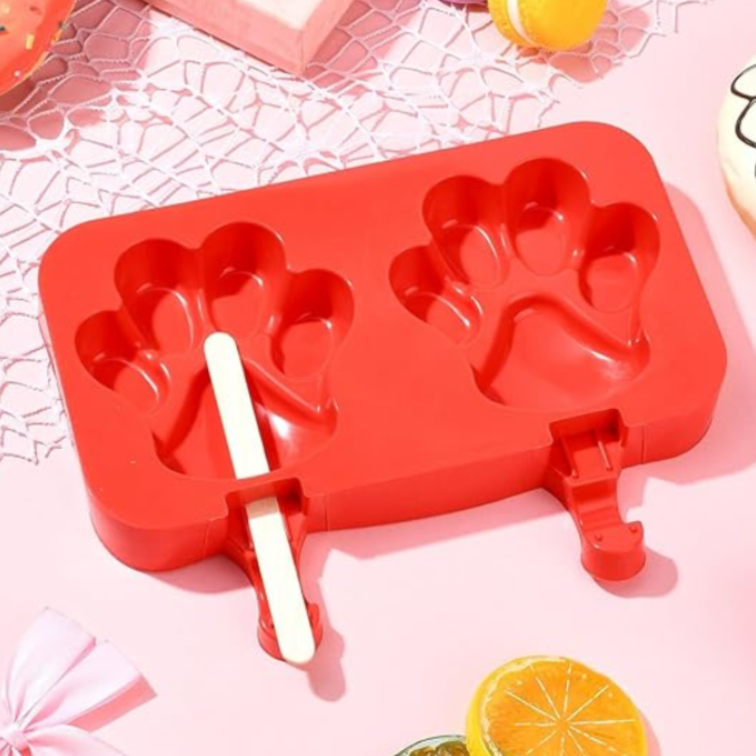 A red silicone popsicle mold with two paw-print shaped cavities and wooden sticks, displayed on a pink surface with decorative macarons, fruit slices, and a gift box.
